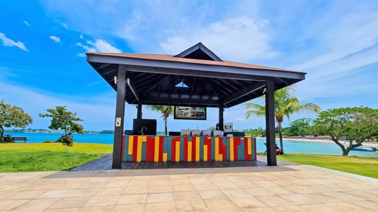 A colourful outdoor jerk hut with a striped red, yellow and blue counter sits beside a grassy lawn overlooking turquoise water. Palm trees and a bright blue sky frame the beachfront setting.