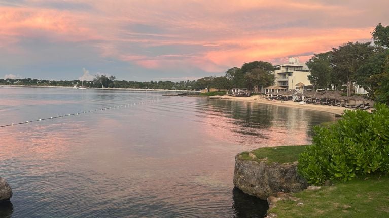 A peaceful beachfront at sunset with calm water reflecting pink and orange clouds. A line of floating buoys stretches across the bay, while resort buildings, trees and palapas line the sandy shore on the right side.