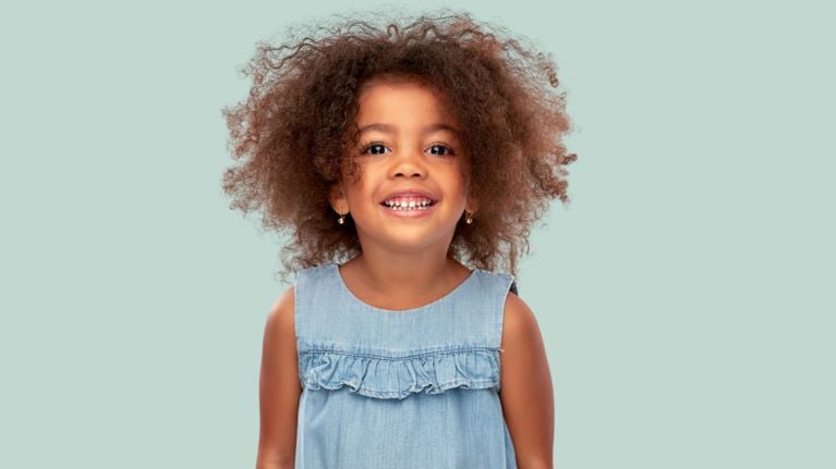 A young child with curly brown hair smiles brightly at the camera. She’s wearing a light denim sleeveless dress with small ruffles on the chest, and simple gold hoop earrings. The background is a soft pastel green.