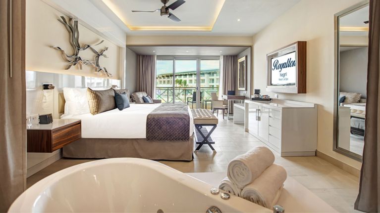 A bright, modern hotel room with a king bed, sofa, large TV and a freestanding bathtub in the foreground. Floor-to-ceiling windows open to a balcony overlooking the resort.