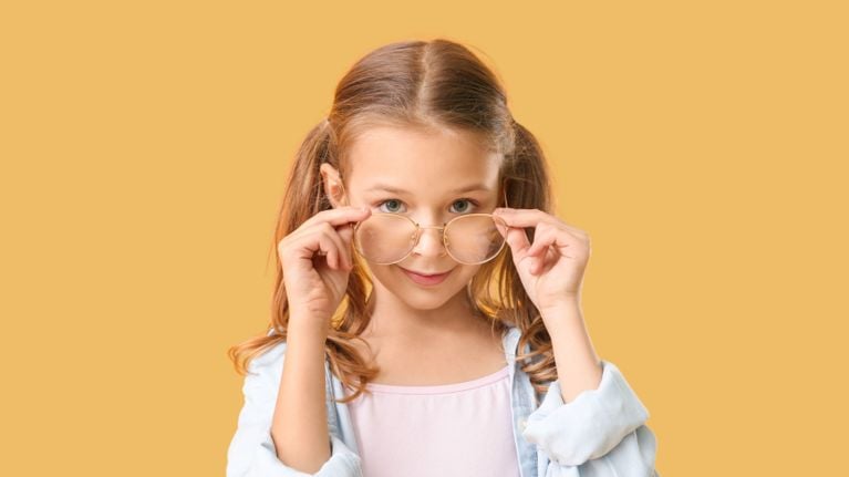 A young girl with light brown hair in pigtails looks over the top of her round eyeglasses with a playful, confident expression. She’s wearing a light denim shirt over a pink top. The background is a warm mustard yellow.