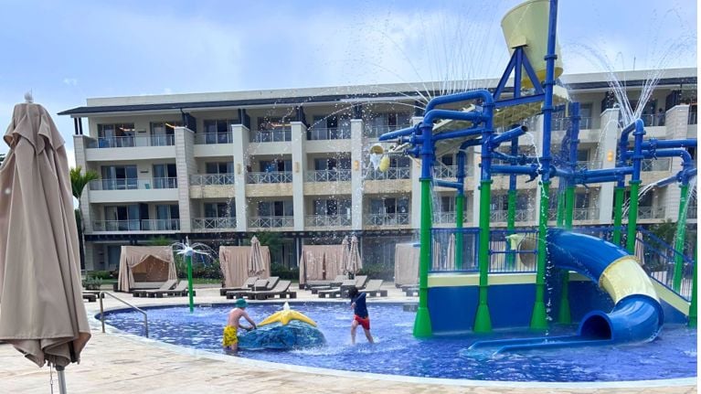 Kids play in the resort’s splash pad, featuring colourful water pipes, sprayers and a small slide. Sun loungers and a multi-storey hotel building sit in the background.