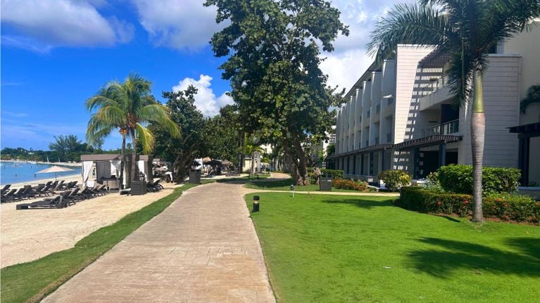 A shaded beachfront walkway lined with palm trees, green lawns and resort buildings on one side, with sandy beach loungers and calm blue water on the other.