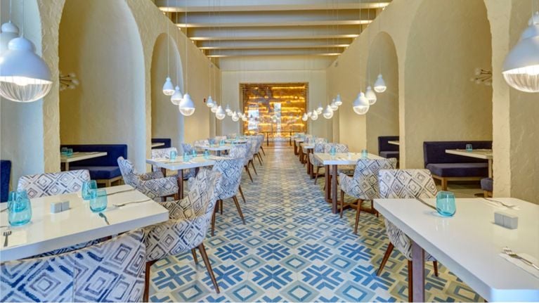 A bright, modern restaurant with patterned blue-and-white tile floors, white tables and cushioned chairs. Rows of pendant lights hang from the ceiling, and arched alcoves with navy booth seating line both sides.