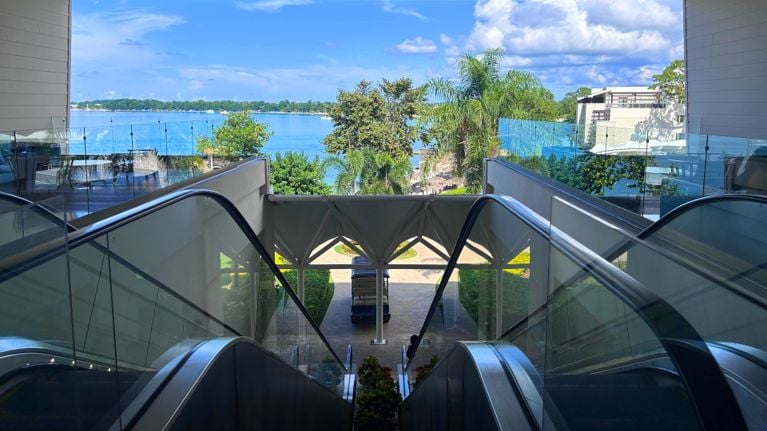 A pair of glass-sided escalators descend from an upper level of the resort, framing a view of lush palm trees, gardens and bright blue ocean under a sunny sky. A small shuttle cart waits at the bottom, with modern resort buildings visible on both sides.
