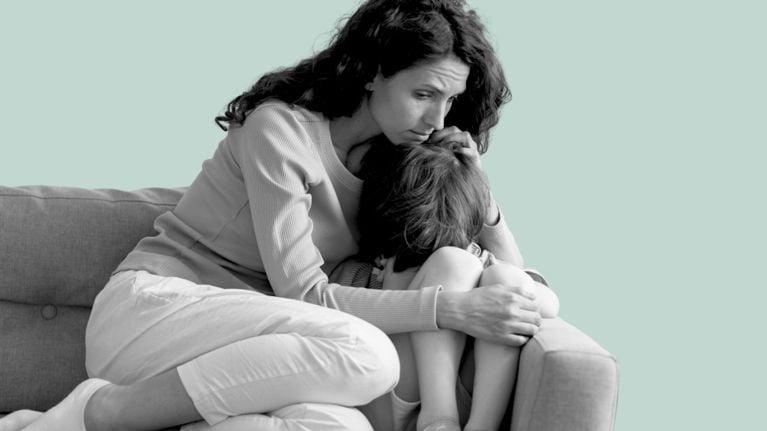 A woman sits on a couch, holding and comforting a child who is curled up beside her, their head tucked into her chest, conveying a quiet, supportive moment.