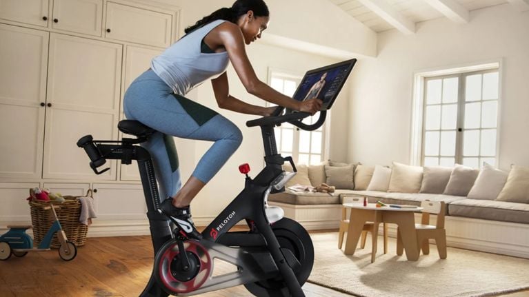 A woman rides a Peloton stationary bike in a bright, cozy living room, watching an instructor on the bike’s screen. A kids’ play area with a small table, chairs, and toys sits nearby, highlighting a family-friendly home workout setup.