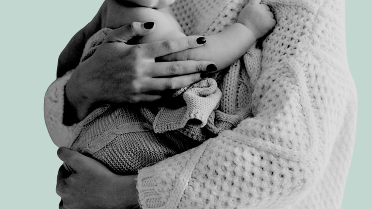 A close-up of a parent wearing a chunky knit sweater, gently cradling a baby wrapped in a soft knit blanket. The parent’s hands rest protectively around the baby, creating a warm, intimate moment against a pale green background.