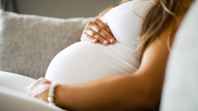 A close-up of a pregnant person reclining on a couch, gently resting their hands on their belly while wearing a white outfit.