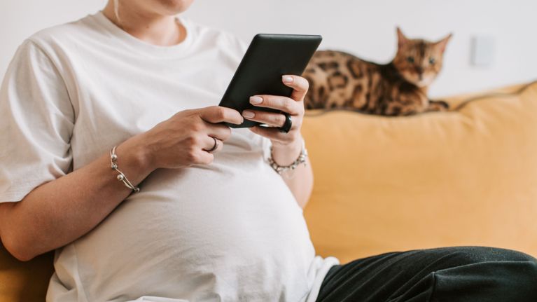 A pregnant person sits on a couch, holding a smartphone while a cat rests on the back of the sofa behind them.