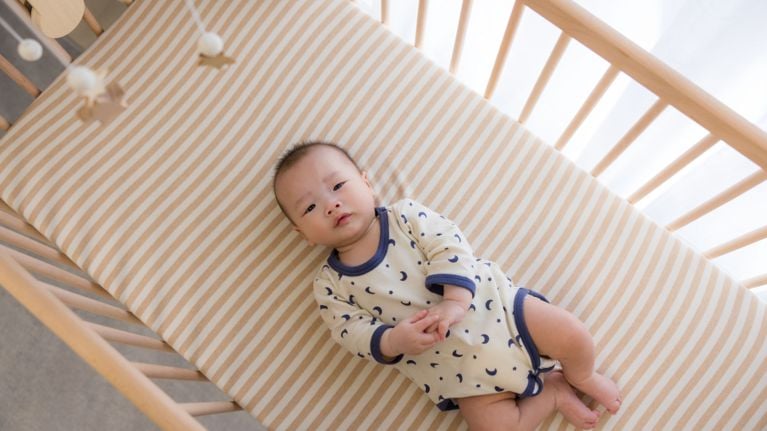 Baby lying awake in a crib on a striped mattress, looking up toward the camera in soft natural light.