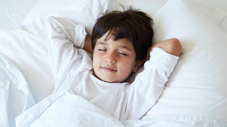 Child sleeping peacefully on white bedding with arms tucked behind their head.