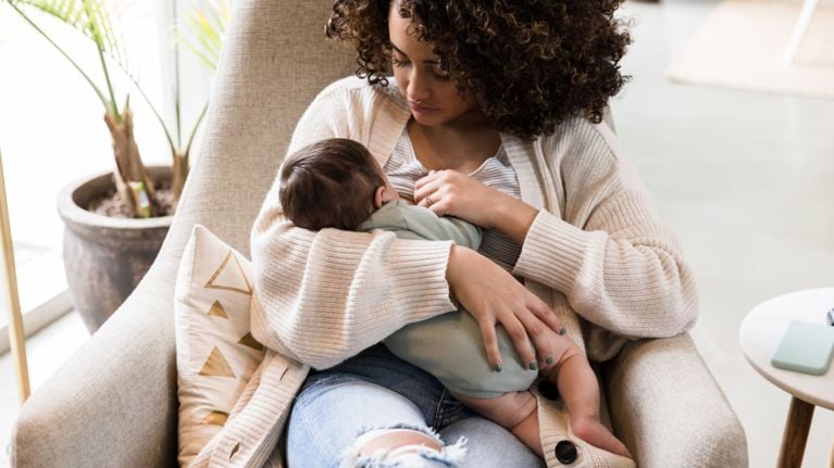 A parent sits in a chair breastfeeding a newborn, cradling the baby close in a calm, sunlit room.