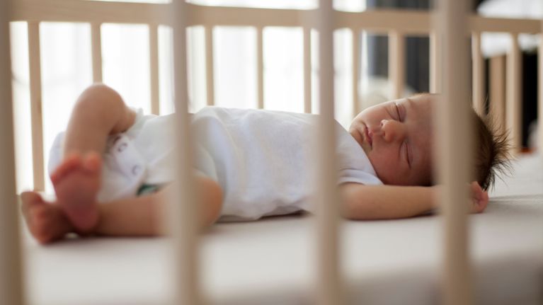 Sleeping baby lying on their back in a crib, viewed through the crib slats in a bright, calm room.