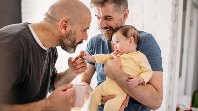 Two dads smile and interact with their baby, one holding the baby while the other reaches out to touch the baby’s hand.