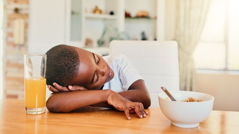 Child resting their head on a kitchen table next to a bowl of cereal and a glass of juice, appearing tired during breakfast.
