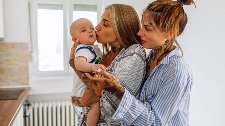 Two parents stand together holding their baby, with one kissing the baby’s cheek in a bright kitchen.