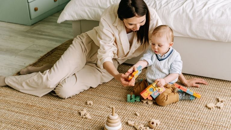 Parent sitting on the floor playing with a baby, both holding colorful wooden puzzle toys on a woven rug in a cozy bedroom.