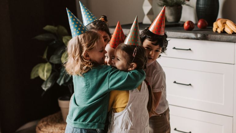 Four diverse young children wearing colorful polka-dot party hats sharing a group hug at a birthday celebration.