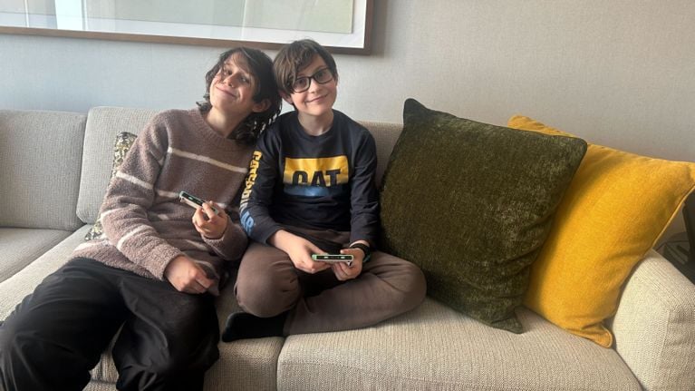 Two young brothers smiling on a grey sofa while holding handheld gaming consoles during a hotel stay.