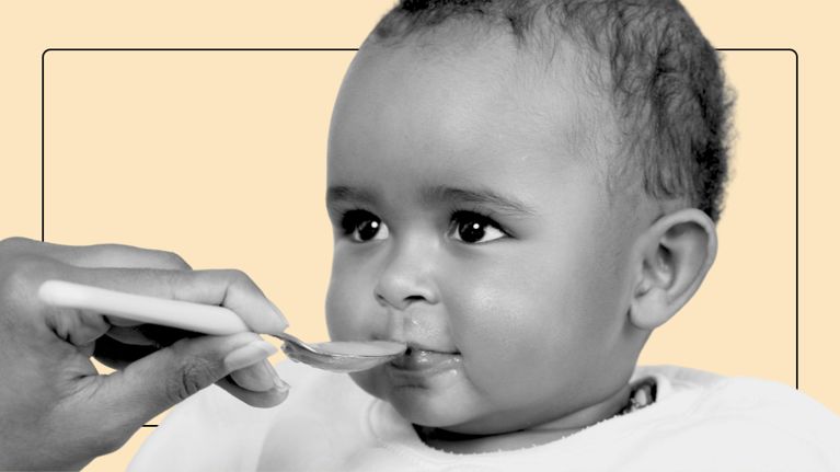 Close-up of a baby being fed with a spoon by an adult hand, shown against a pale background.
