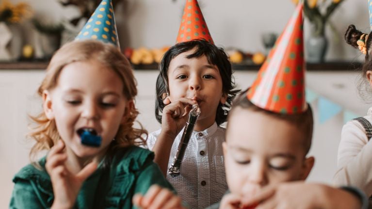 A young boy with dark curly hair wearing a red polka-dot party hat and blowing a silver tinsel party horn.