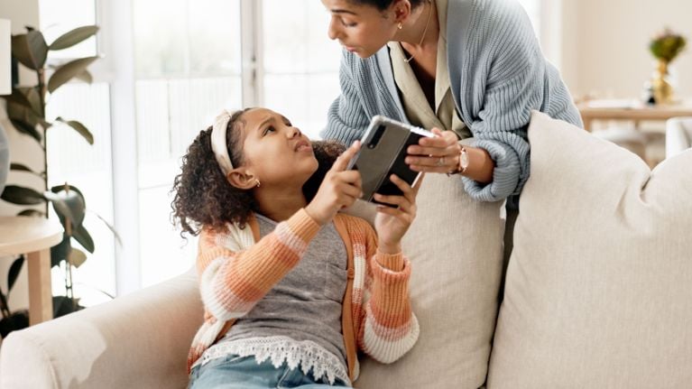 A woman leans over a couch where a young girl sits holding a tablet. The woman is pulling the tablet out of the girl's hands.