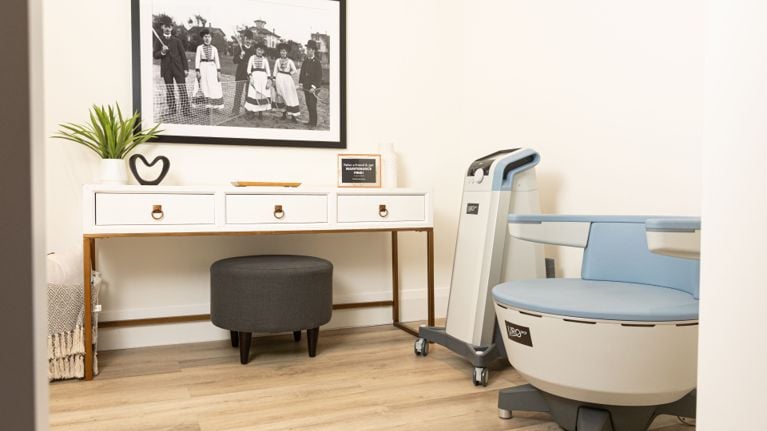 A clean, brightly lit clinical room designed for pelvic floor therapy. The scene includes the UroSpot chair and console, a grey ottoman, and a white console table decorated with a vintage photo of tennis players, a plant, and a referral sign.