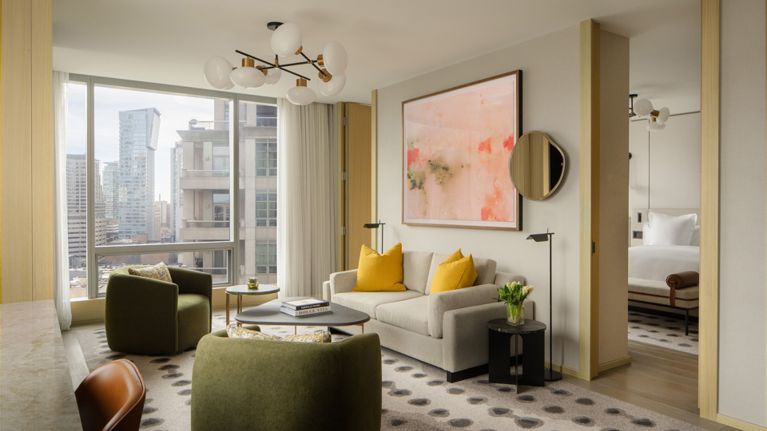 A modern, sunlit luxury hotel living room with floor-to-ceiling windows overlooking a city skyline.