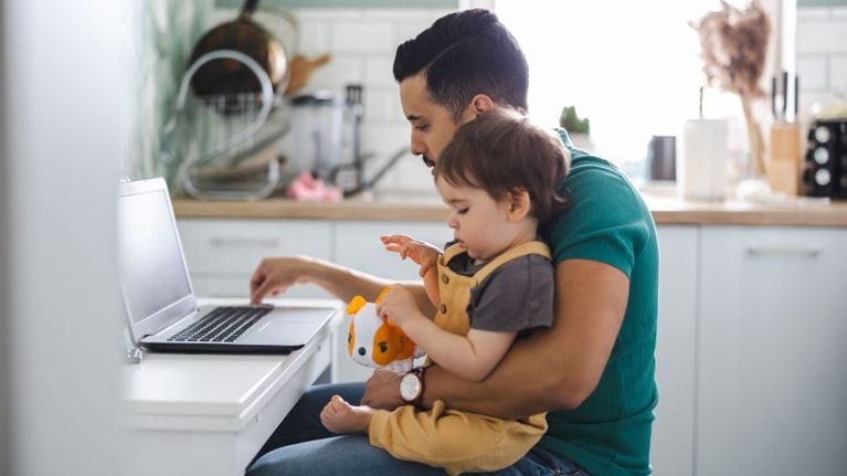 An adult man with short dark hair and a mustache sits at a white desk in a kitchen, working on a laptop while holding a young child on his lap. The man is wearing a teal t-shirt and a watch, appearing focused on the screen. The child, dressed in yellow overalls, sits facing sideways and plays with a small orange and white toy dog. The background shows a bright, modern kitchen with white cabinetry and wooden countertops.