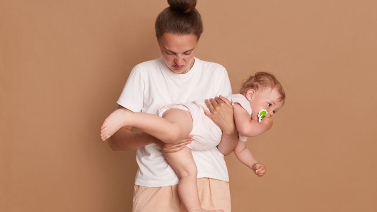 A relatable parenting moment showing a mother’s "stinky face" expression of disgust as she carries her baby, who has a green pacifier. The image perfectly captures the messy, unglamorous side of diaper changes and toddler care.