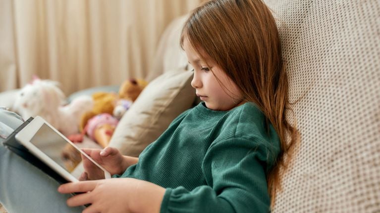 Young girl with long brown hair sitting on a sofa using a tablet for educational screen time.