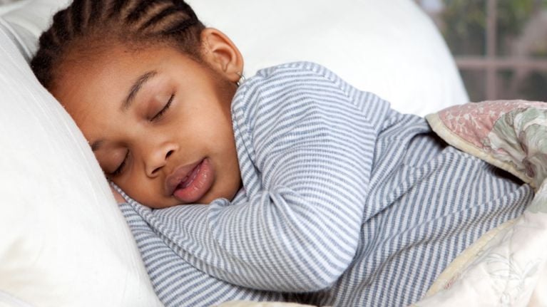 A young girl with braided hair sleeping soundly on a white pillow, wearing striped pajamas under a quilt.