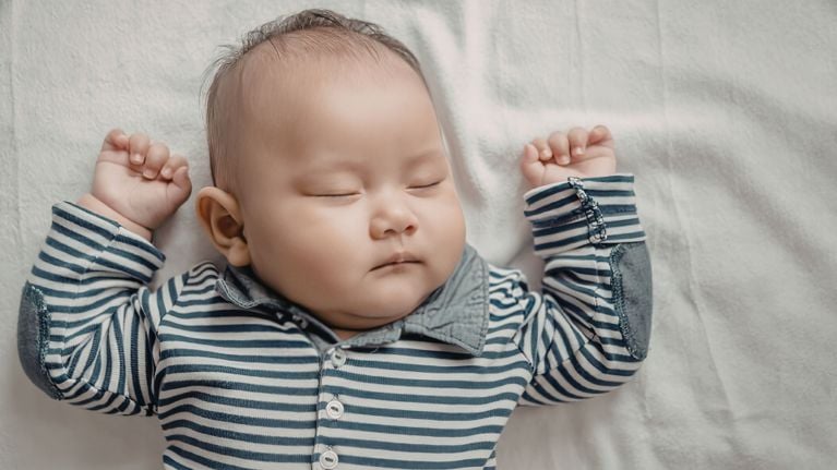 Chubby baby with dark hair sleeping on a white sheet in a blue and white striped shirt.