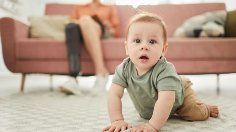 A curious baby crawls on a rug toward the camera while a parent with a prosthetic leg sits on a pink sofa.