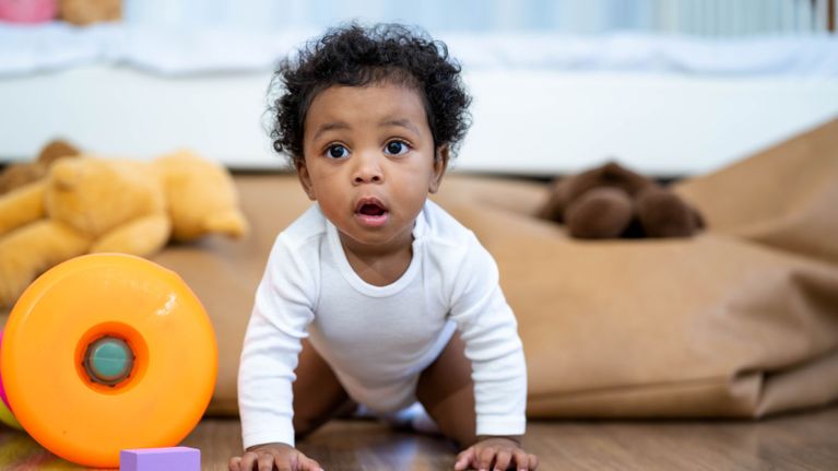 A wide-eyed baby with curly hair crawls on a wooden floor toward a bright orange toy.