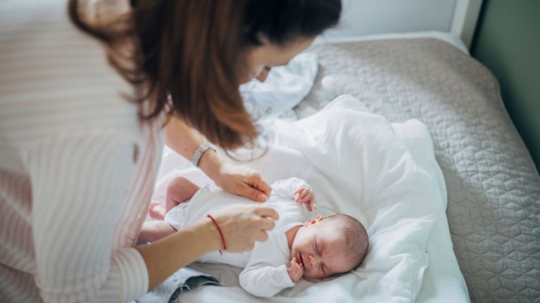A mother gently leans over her newborn baby on a bed, attempting to dress them in a white long-sleeved onesie while the infant cries. The scene captures the realistic, everyday challenges of newborn care and the focus required during diaper and outfit changes.