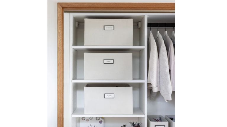 A well-organized white closet featuring three structured fabric bins labeled "Athleisure," "School," and "Pajamas." To the right, children's sweaters and shirts are neatly hung on matching white hangers.