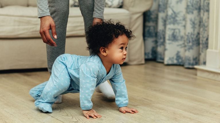 A curious baby crawls on a rug toward the camera while a parent with a prosthetic leg sits on a pink sofa.