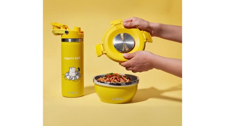 A bright yellow lunch set including a stainless steel lined bowl filled with spaghetti and a matching "Thirsty One!" water bottle. A person is shown lifting the lid off the bowl to reveal the meal inside.