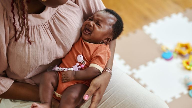A mother holds her distressed infant, who is crying while wearing a rust-orange ribbed onesie with a beaded pacifier clip. The close-up shot emphasizes the emotional connection and the common parenting experience of soothing a fussy baby.