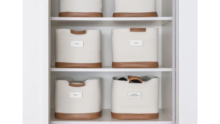 White coiled rope baskets with tan leather bases and handles are arranged on white shelves. Each basket features a white label identifying contents like "Swim" and "Winter Accessories," showcasing a tidy system for seasonal gear.