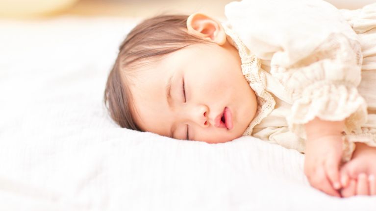 Close-up of a baby with dark hair sleeping in a white lace-collared vintage-style dress.
