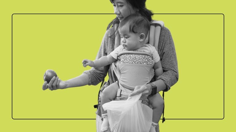 A mother wearing a baby carrier inspects an apple while holding a plastic grocery bag.