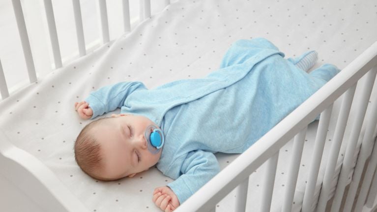 Infant sleeping in a white crib wearing blue footie pajamas with a blue pacifier.