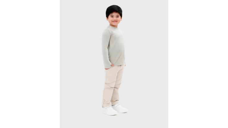 A child stands comfortably in UPF clothing, featuring a shirt. He is wearing white sneakers and looking toward the camera with a cheerful expression.