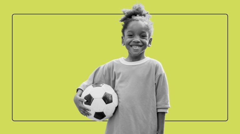 A smiling young girl in a jersey proudly holds a soccer ball under her arm.