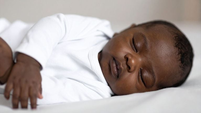 Baby in a white long-sleeve onesie sleeping peacefully on a white bed.