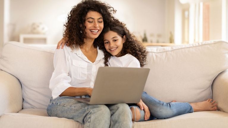 A smiling mother and young daughter with curly hair sit on a couch together looking at a laptop computer.