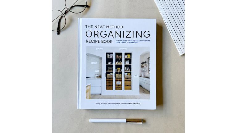 A white hardcover book titled "The Neat Method Organizing Recipe Book" sits on a tan surface, surrounded by lifestyle accessories including wire-rimmed glasses, a polka-dot notebook, and a sleek white pen. The cover features a photo of a perfectly organized pantry with labels, promoting "70 simple projects to take your home from chaos to composed."